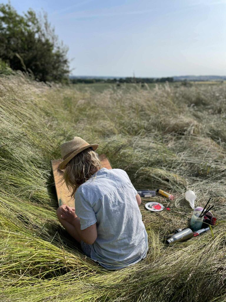 Woman in a hat painting while sitting in tall grass